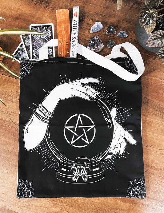 Pictured is a large canvas tote bag featuring an image of a black crystal ball with hands around it and a pentagram in the middle of the crystal ball. Black Crystal Ball & Pentacle Tote Bag