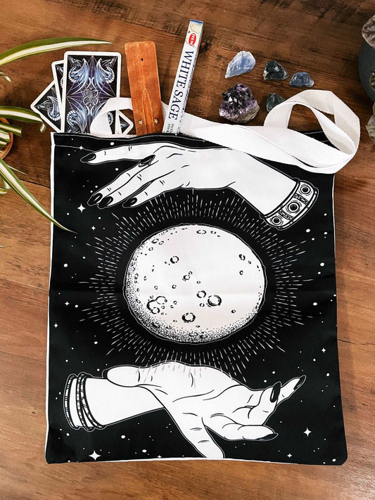 Pictured is a large canvas tote bag featuring an image of two hands around an image of a moon. Black Moon & Hands Tote Bag