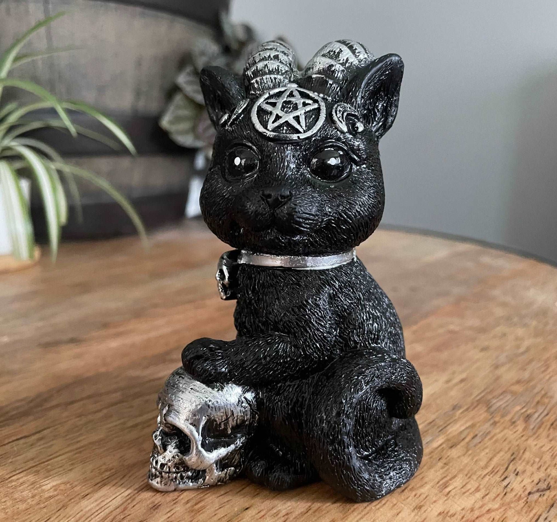 Pictured is a resin statue of a black cat with horns and a pentagram on its forehead with a paw on a silver skull. Demon Cat Figurine