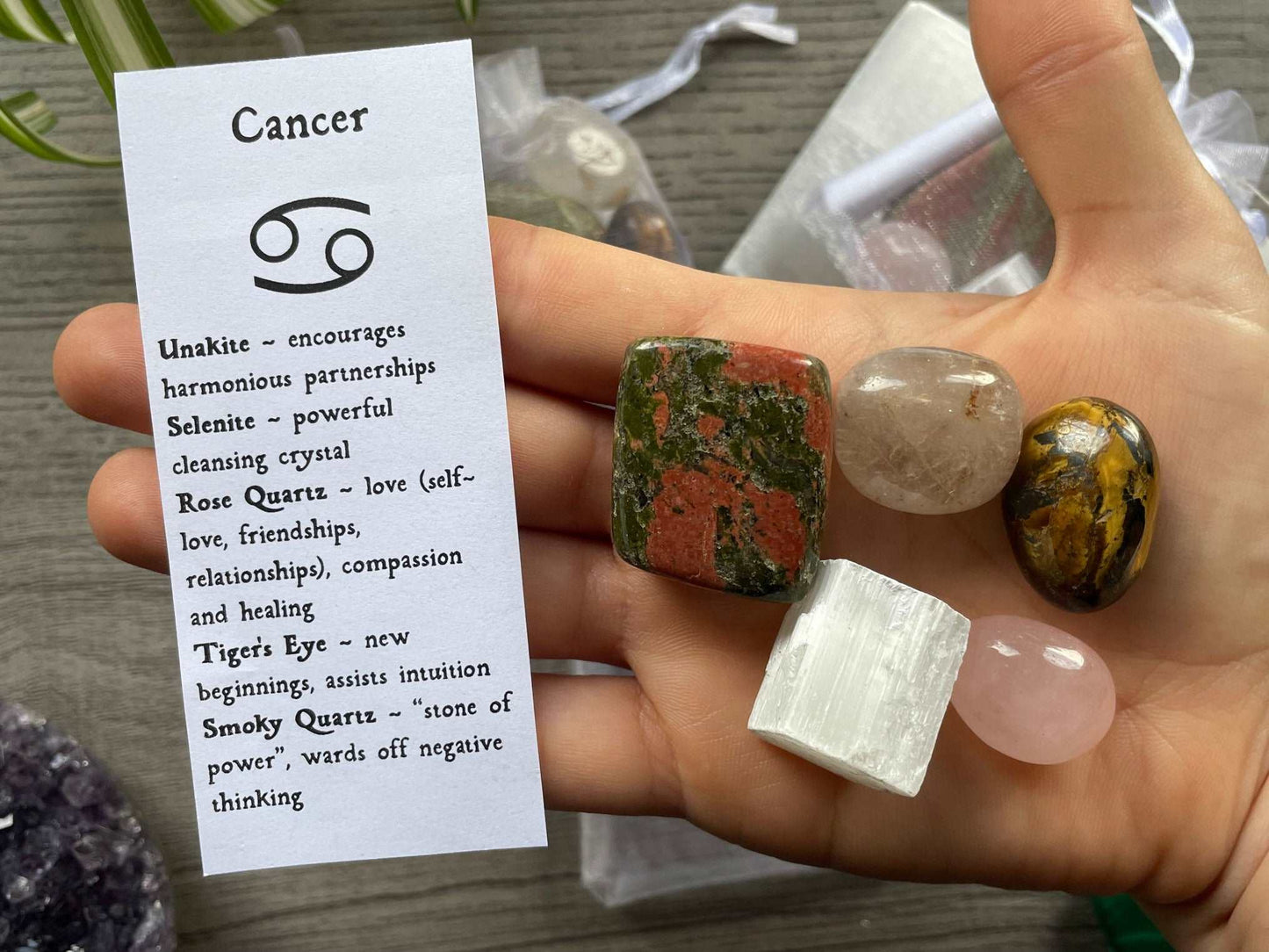 An image of a collection of tumbled stones in an organza bag. To the side is a piece of paper that describes what each tumbled stone's metaphysical properties are. CANCER Zodiac Tumbled Crystal Bag crystals