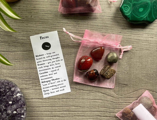 An image of a collection of tumbled stones in an organza bag. To the side is a piece of paper that describes what each tumbled stone's metaphysical properties are. FOCUS Tumbled Crystal Bag