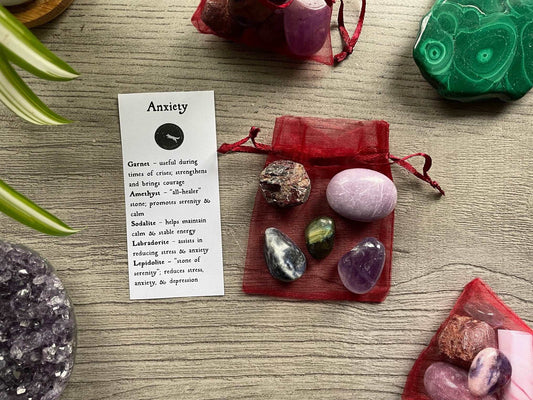 An image of a collection of tumbled stones in an organza bag. To the side is a piece of paper that describes what each tumbled stone's metaphysical properties are. ANXIETY Tumbled Crystal Bag