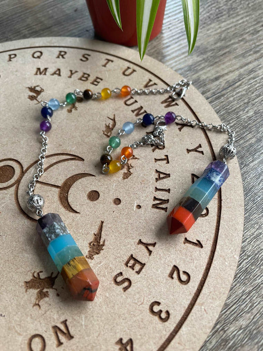 A multicoloured crystal pendulum sits atop a wooden divination board. Chakra Crystals Pendulum 