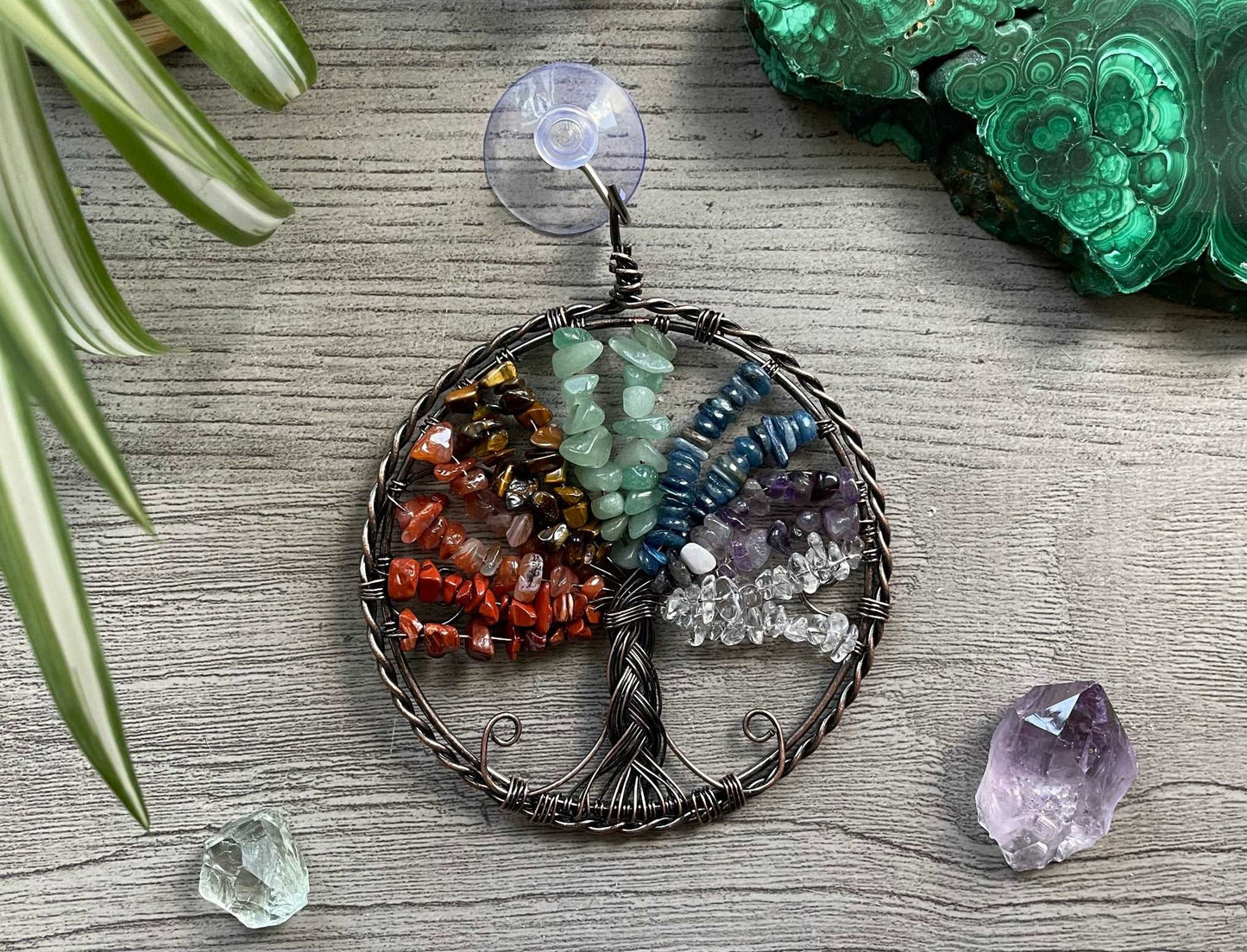 An image of a window art piece with a suction cup. The art is a wire-wrapped tree of life and the leaves on the tree are polished chips of green aventurine, red jasper, carnelian, tiger's eye, aquamarine, amethyst, and clear quartz. Chakra Crystal Gemstones Wire-Wrapped Tree of Life Window Decoration