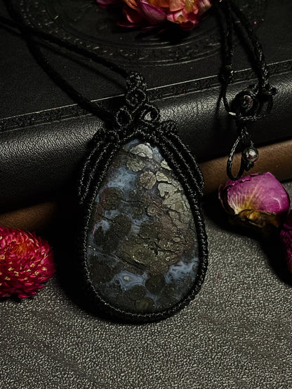 Pictured is a marcasite in agate cabochon wrapped in macrame thread. A gothic book and flowers are nearby. Marcasite in Agate Macramé Necklace (Twisted Nightshade Jewellery) - The Wandering Fox Emporium, Your Crystal Store front