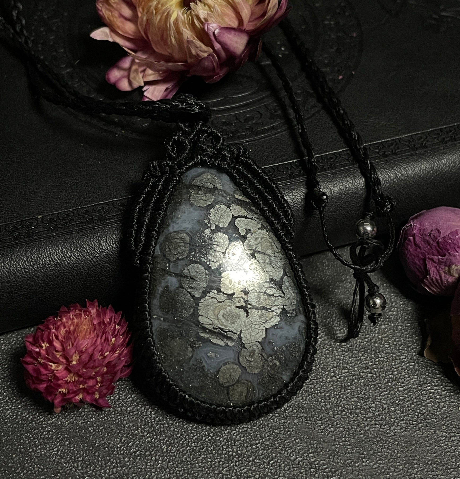 Pictured is a marcasite in agate cabochon wrapped in macrame thread. A gothic book and flowers are nearby. Marcasite in Agate Macramé Necklace (Twisted Nightshade Jewellery) - The Wandering Fox Emporium, Your Crystal Store