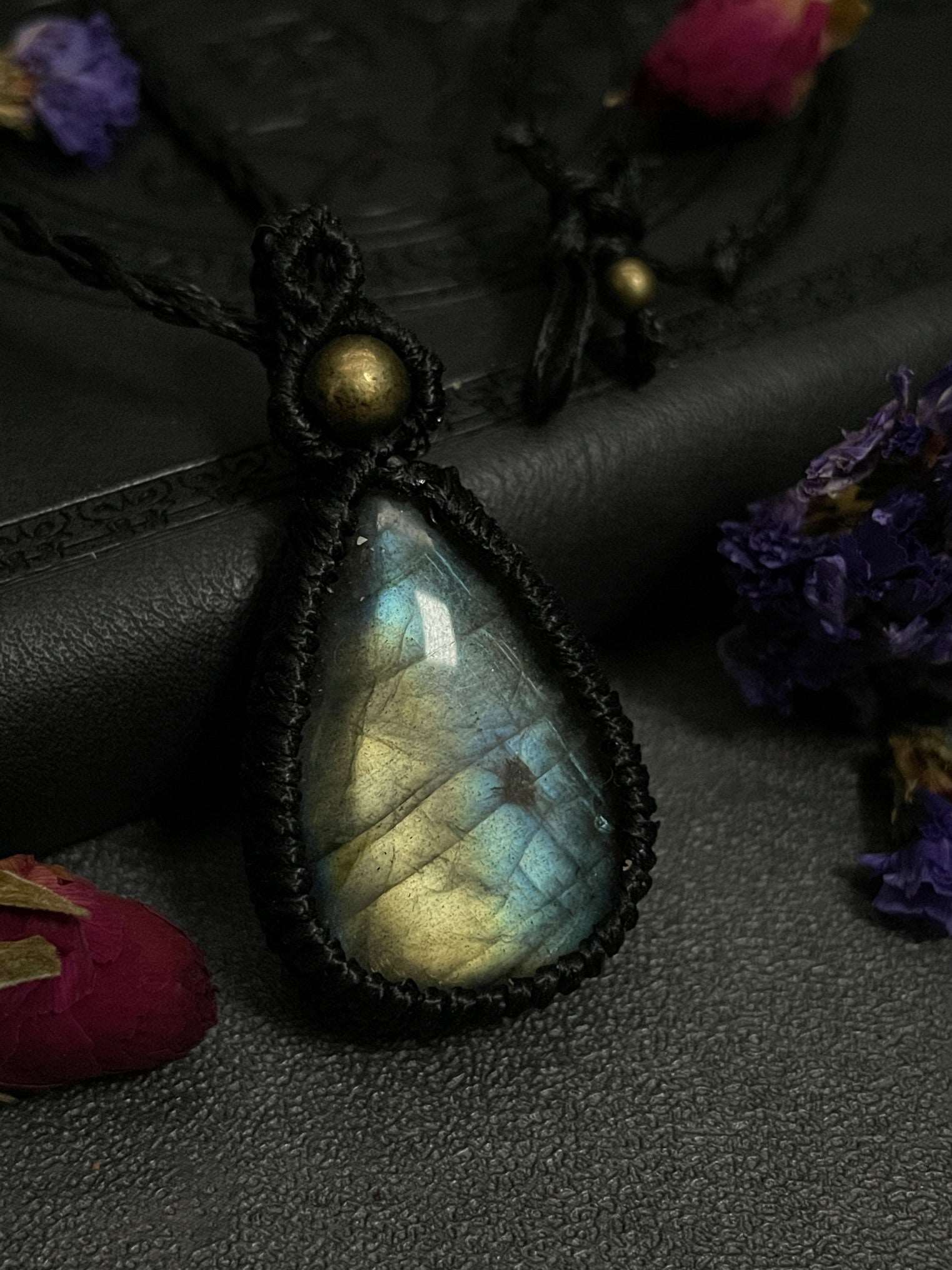 Pictured is a labradorite cabochon wrapped in macrame thread. A gothic book and flowers are nearby. Blue/Gold Labradorite Teardrop Macramé Necklace (Twisted Nightshade Jewellery) - The Wandering Fox Emporium, Your Crystal Store