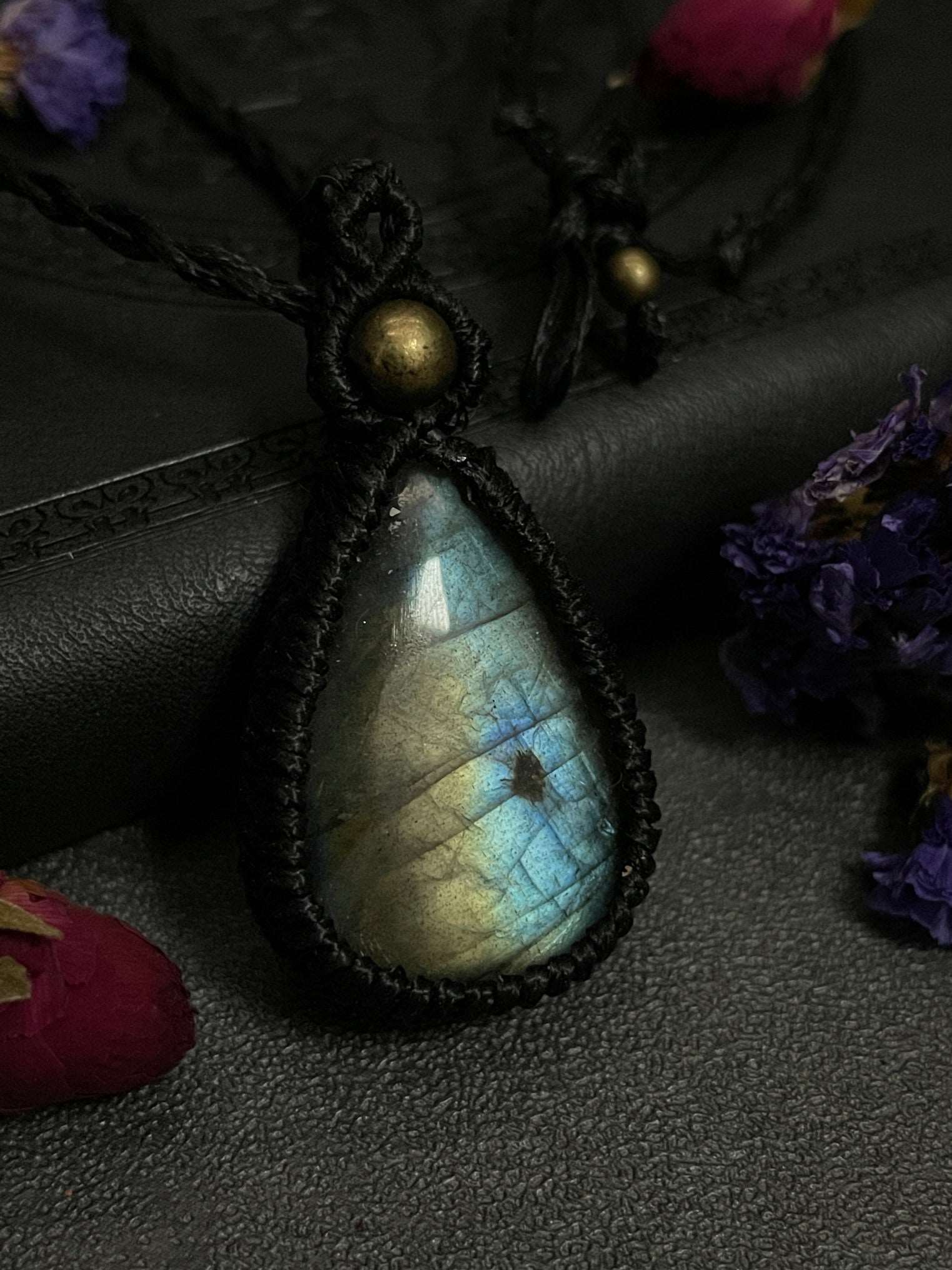 Pictured is a labradorite cabochon wrapped in macrame thread. A gothic book and flowers are nearby. Blue/Gold Labradorite Teardrop Macramé Necklace (Twisted Nightshade Jewellery) - The Wandering Fox Emporium, Your Crystal Store close up