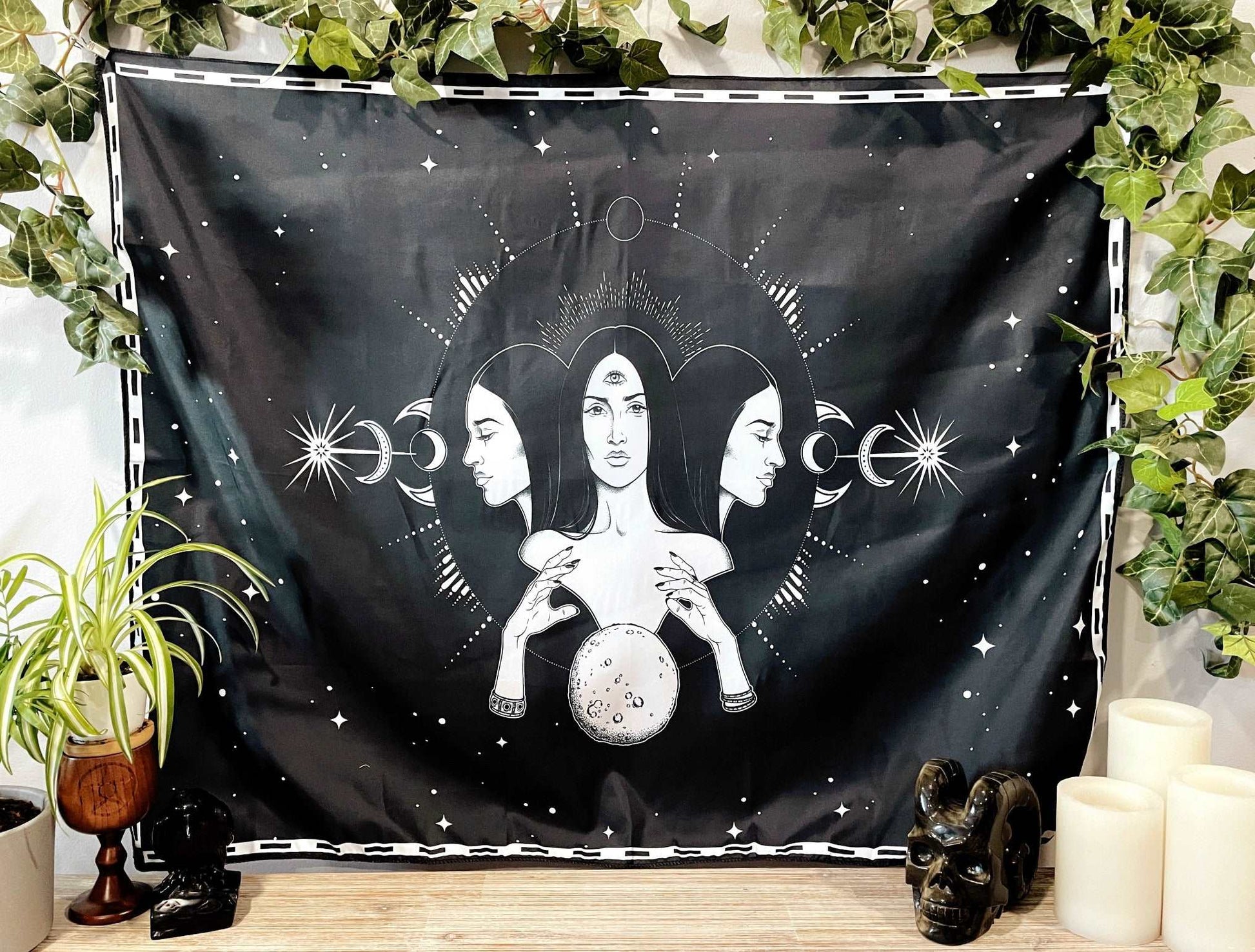 Pictured is a large wall tapestry with a black background and three women representing three goddesses or Hecate printed in white on it. Goddesses & Moon Wall Tapestry