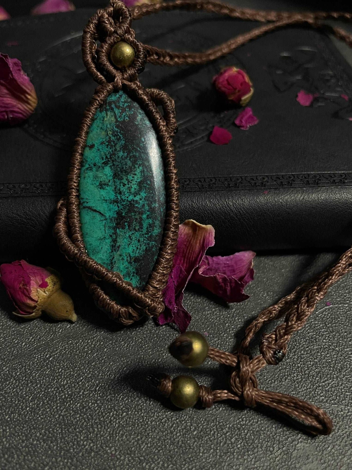 Pictured is a chrysocolla cabochon wrapped in macrame thread. A gothic book and flowers are nearby. Chrysocolla Macramé Necklace (Twisted Nightshade Jewellery) - The Wandering Fox Emporium, Your Crystal Store close up