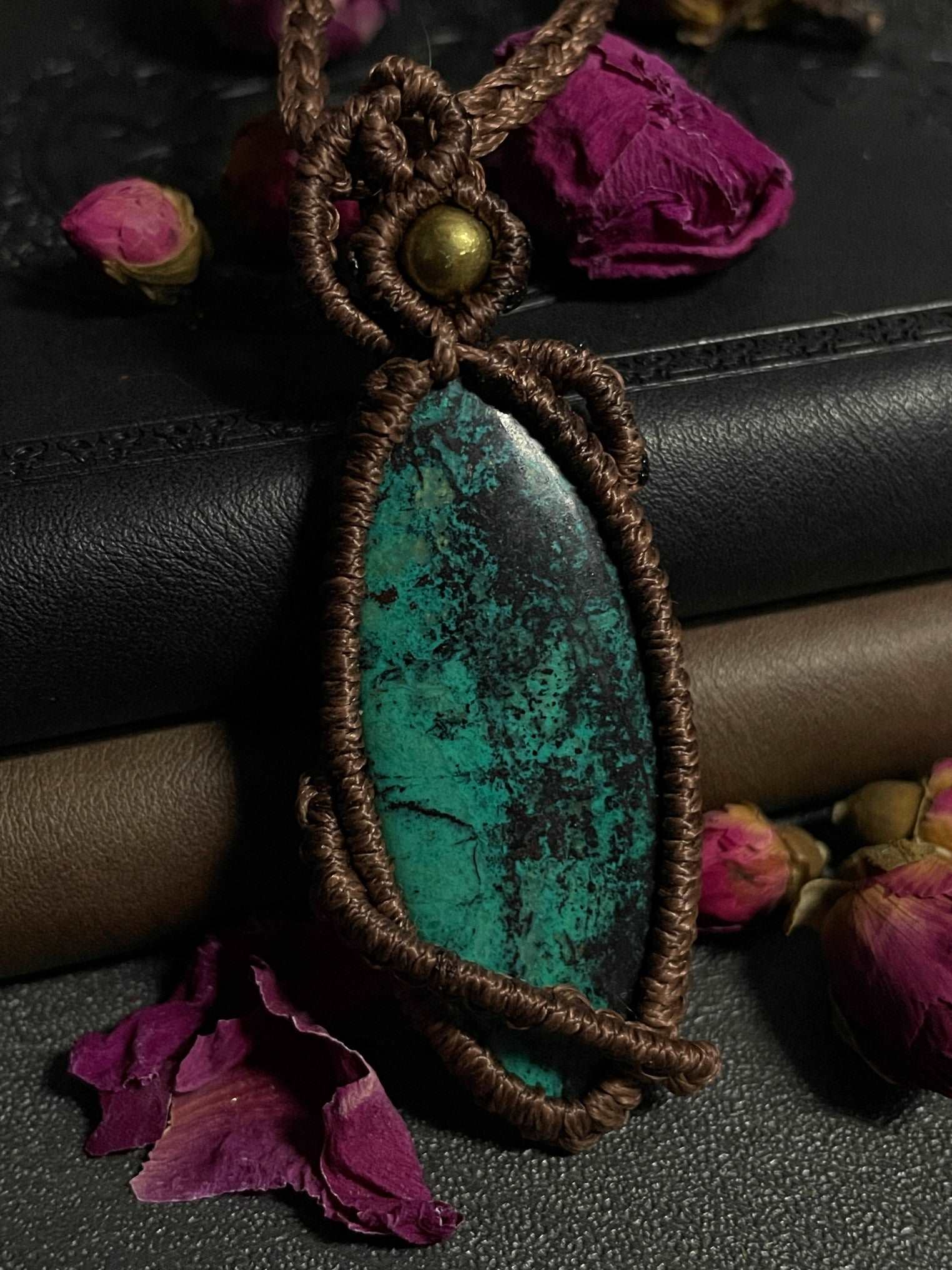 Pictured is a chrysocolla cabochon wrapped in macrame thread. A gothic book and flowers are nearby. Chrysocolla Macramé Necklace (Twisted Nightshade Jewellery) - The Wandering Fox Emporium, Your Crystal Store