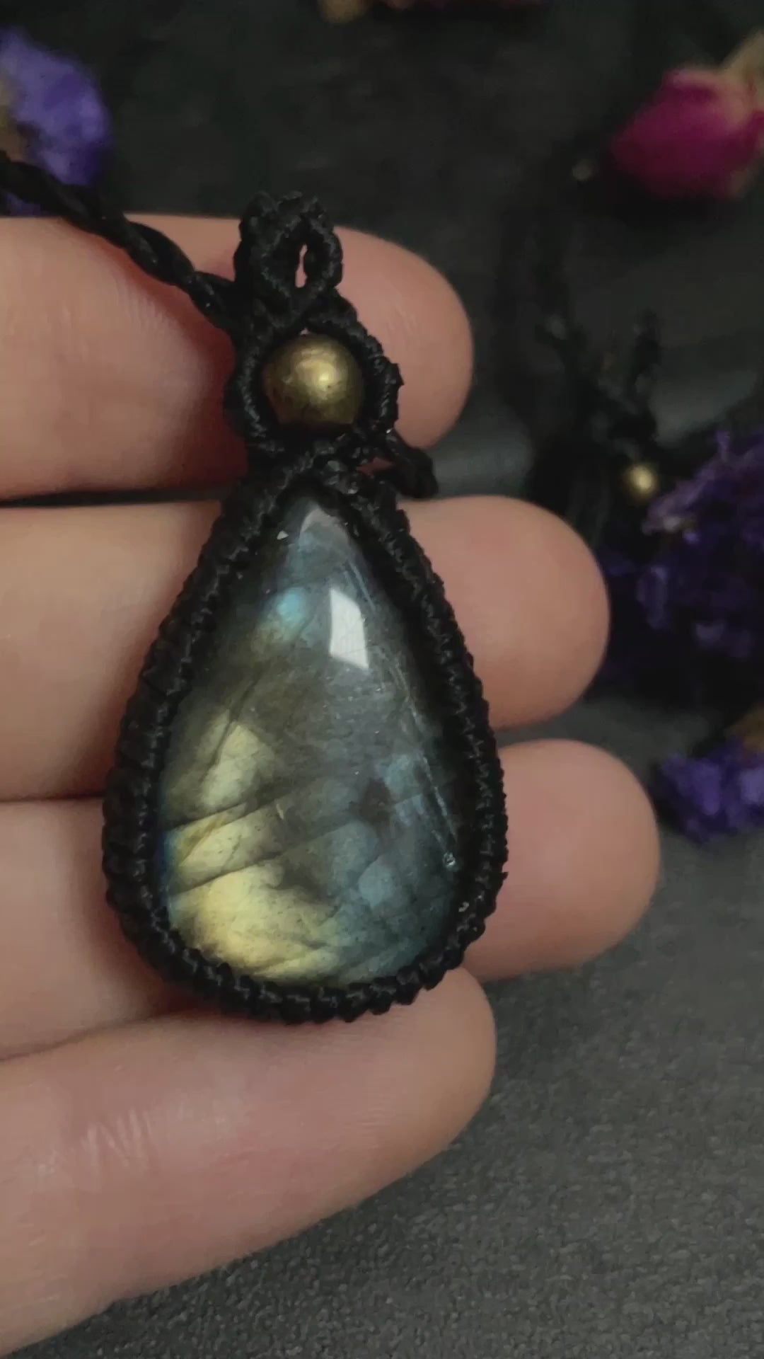 Pictured is a labradorite cabochon wrapped in macrame thread. A gothic book and flowers are nearby. Blue/Gold Labradorite Teardrop Macramé Necklace (Twisted Nightshade Jewellery) - The Wandering Fox Emporium, Your Crystal Store video