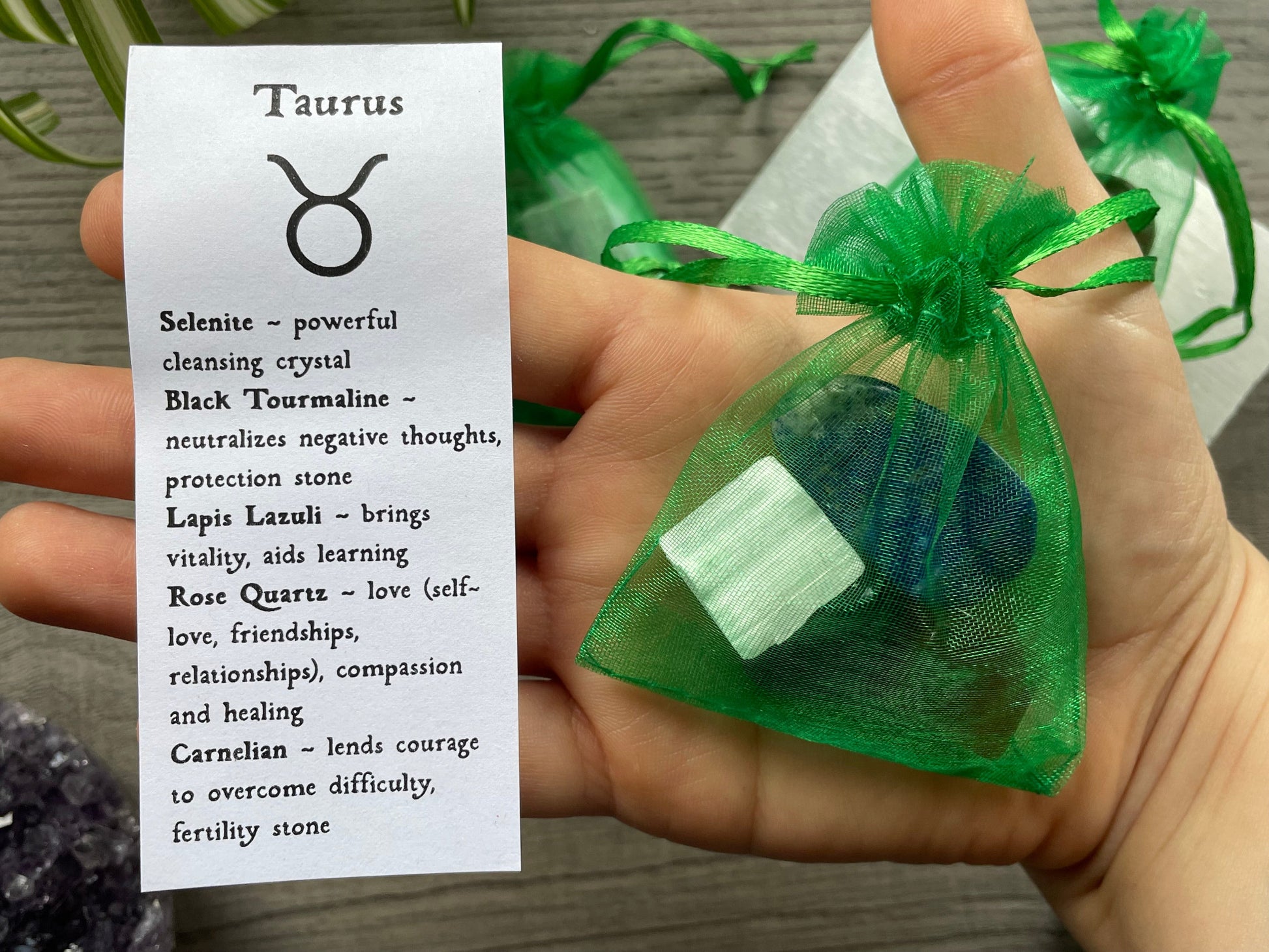 An image of a collection of tumbled stones in an organza bag. To the side is a piece of paper that describes what each tumbled stone's metaphysical properties are. TAURUS Zodiac Tumbled Crystal Bag front