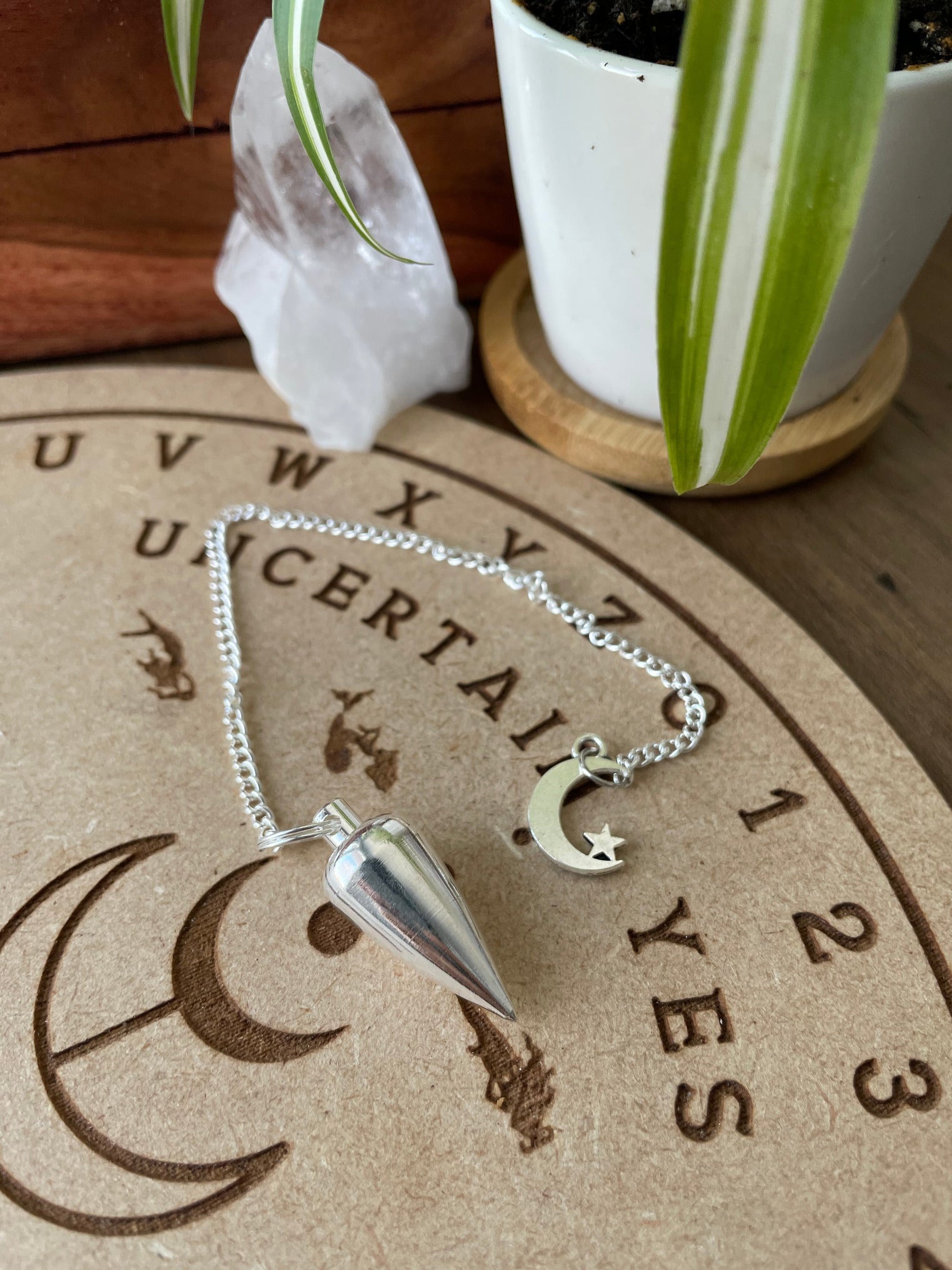 A silver coloured pendulum with a crescent moon and star sits atop a wooden divination board. Metal Moon & Star Pendulum 