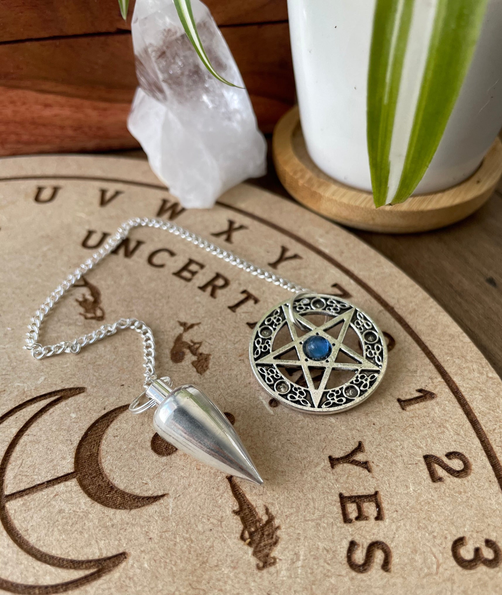 A silver coloured pendulum with a pentagram sits atop a wooden divination board. Metal Pentacle Pendulum