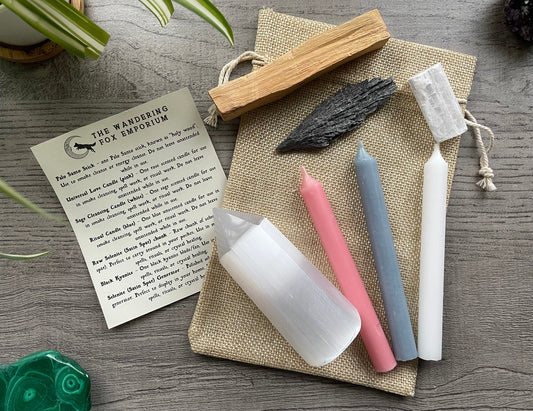 Pictured is a protection and cleansing starter set with candles, selenite, kyanite, palo alto, and a piece of paper explaining how to use the items next to it. Protection & Cleansing Starter Set