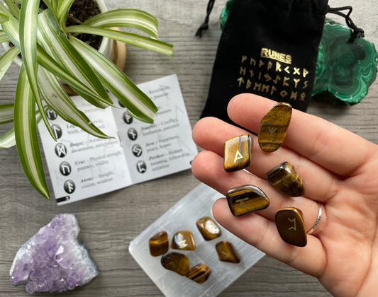 Pictured is a rune set made of tiger's eye tumbled stones. A black velvet bag and instructions sit nearby, Tiger's Eye Mini Rune Set (24 runes)