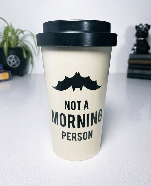 An image of a white travel mug with a black bat design and the phrase "not a morning person" written in bold black letters. The mug is tall and cylindrical with a black plastic lid that has a sipping hole and a sliding tab to open and close it.  "Not a Morning Person" Eco Travel Mug front