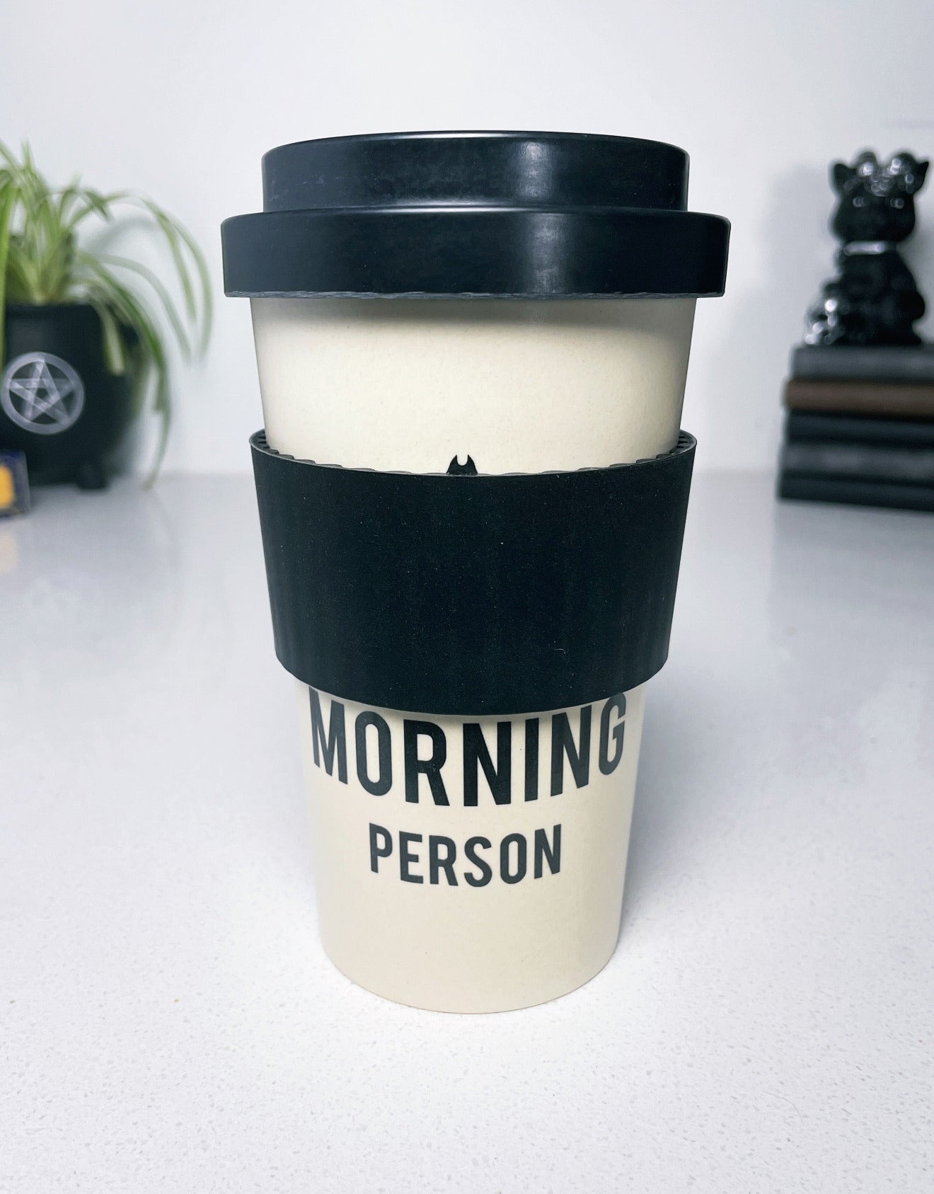 An image of a white travel mug with a black bat design and the phrase "not a morning person" written in bold black letters. The mug is tall and cylindrical with a black plastic lid that has a sipping hole and a sliding tab to open and close it. "Not a Morning Person" Eco Travel Mug sleeve