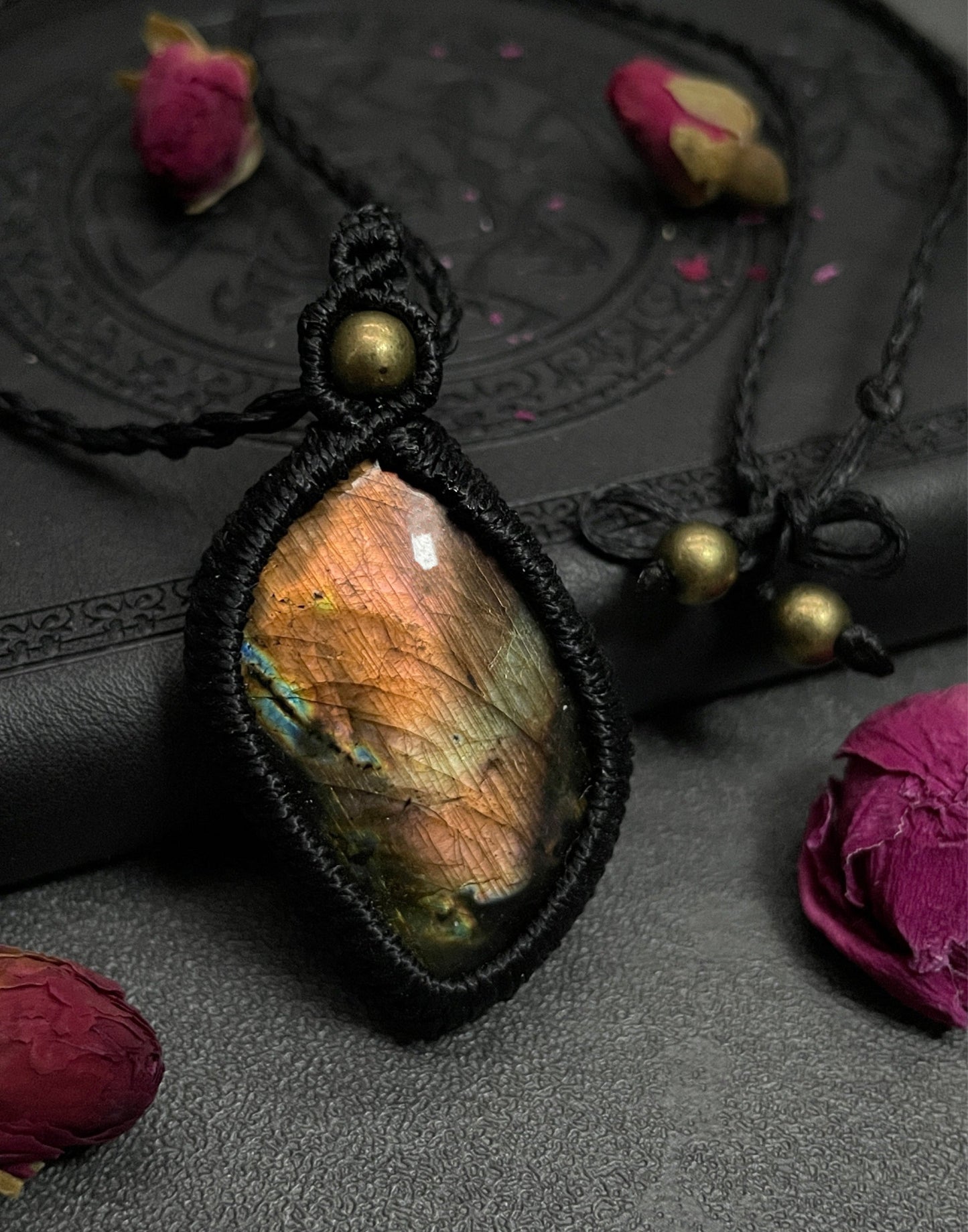 Pictured is a labradorite cabochon wrapped in macrame thread. A gothic book and flowers are nearby. Sunset Labradorite Macramé Necklace (Twisted Nightshade Jewellery) - The Wandering Fox Emporium, Your Crystal Store