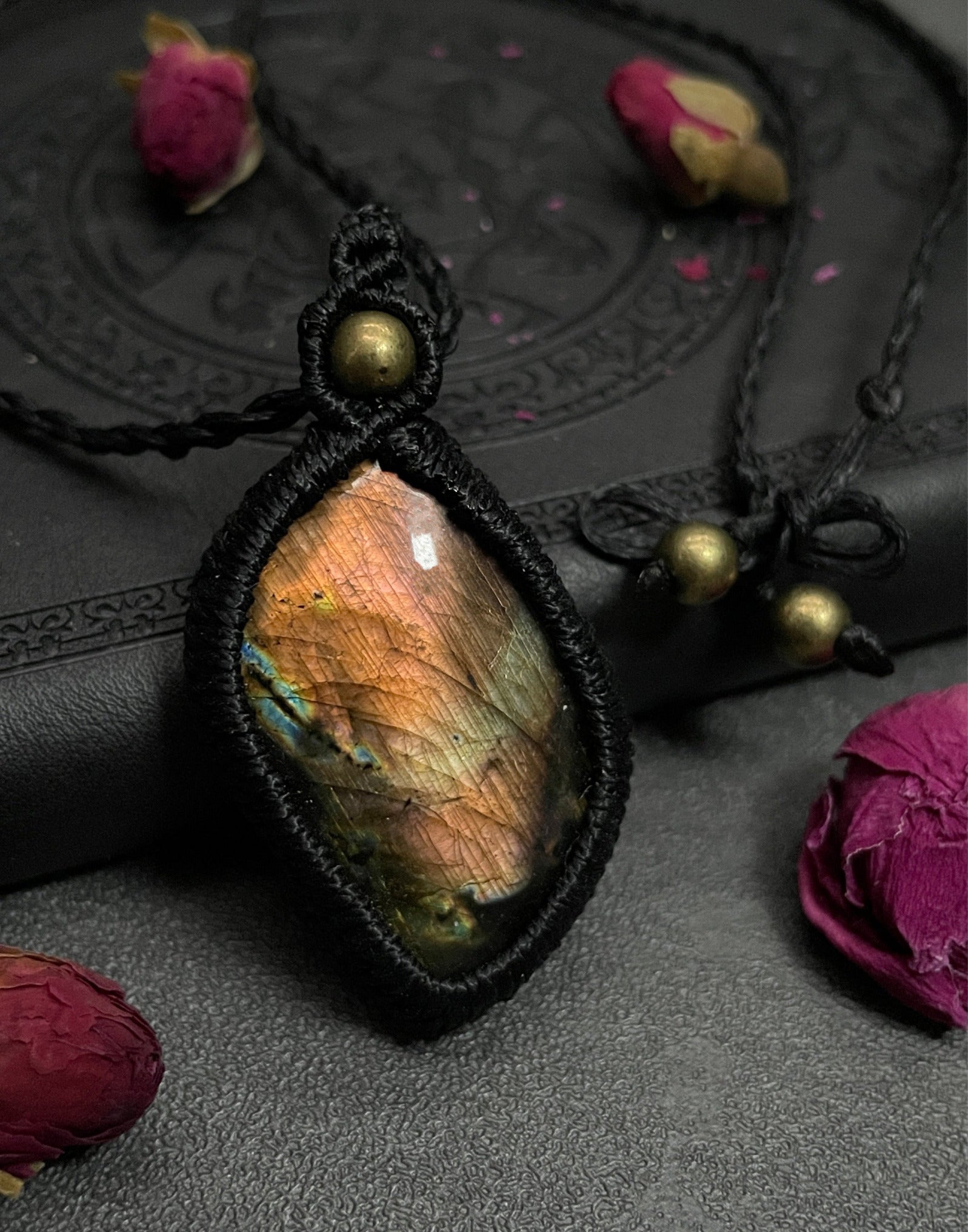 Pictured is a labradorite cabochon wrapped in macrame thread. A gothic book and flowers are nearby. Sunset Labradorite Macramé Necklace (Twisted Nightshade Jewellery) - The Wandering Fox Emporium, Your Crystal Store