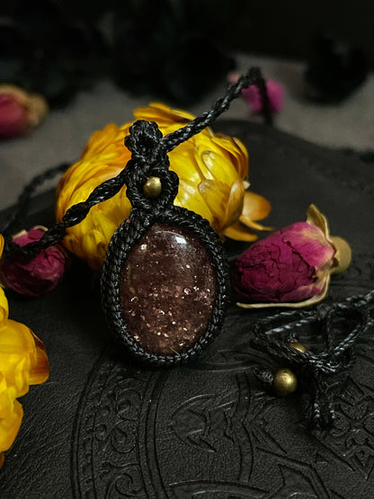 Pictured is a strawberry quartz cabochon wrapped in macrame thread. A gothic book and flowers are nearby. Strawberry Quartz Macramé Necklace (Twisted Nightshade Jewellery) - The Wandering Fox Emporium, Your Crystal Store