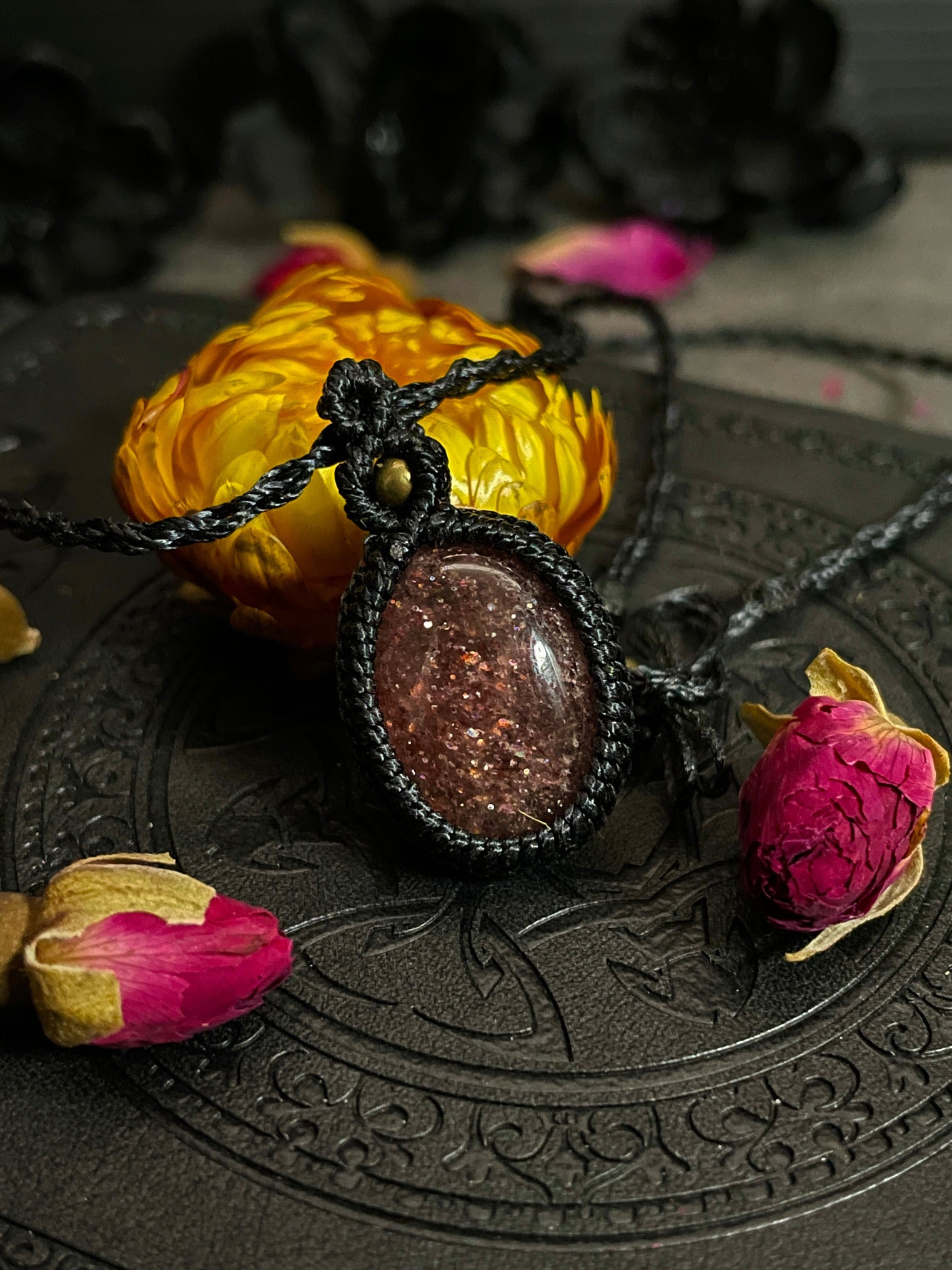 Pictured is a strawberry quartz cabochon wrapped in macrame thread. A gothic book and flowers are nearby. Strawberry Quartz Macramé Necklace (Twisted Nightshade Jewellery) - The Wandering Fox Emporium, Your Crystal Store front