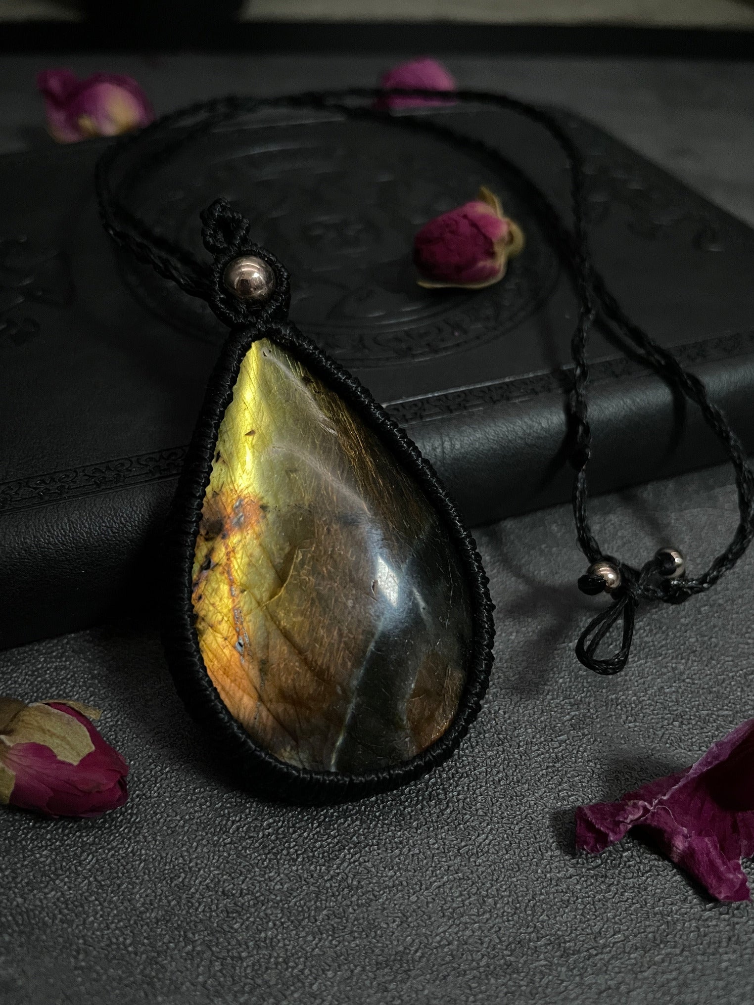 Pictured is a labradorite cabochon wrapped in macrame thread. A gothic book and flowers are nearby. Labradorite Teardrop Macramé Necklace (Twisted Nightshade Jewellery) - The Wandering Fox Emporium, Your Crystal Store front