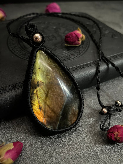 Pictured is a labradorite cabochon wrapped in macrame thread. A gothic book and flowers are nearby. Labradorite Teardrop Macramé Necklace (Twisted Nightshade Jewellery) - The Wandering Fox Emporium, Your Crystal Store close up