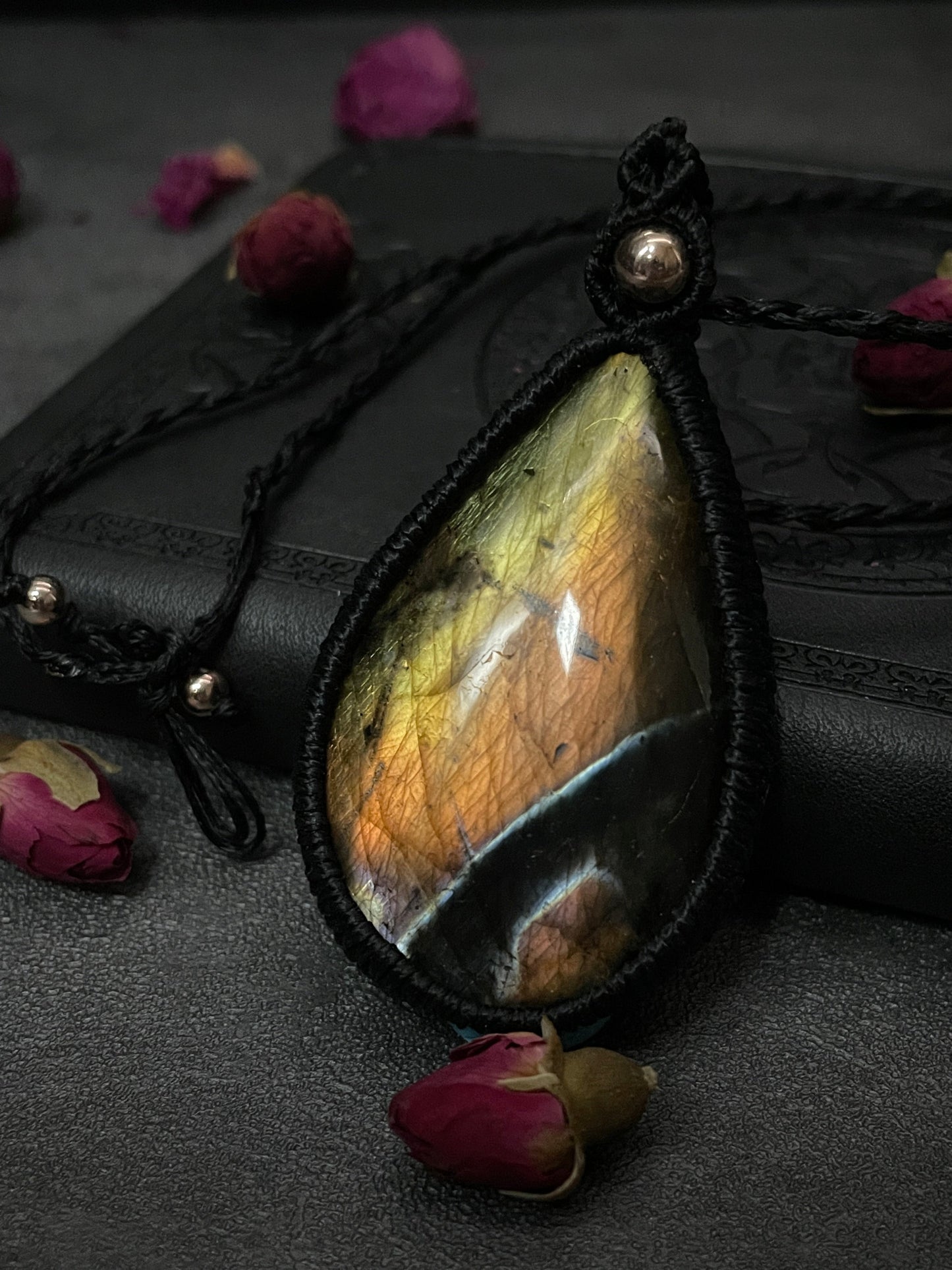 Pictured is a labradorite cabochon wrapped in macrame thread. A gothic book and flowers are nearby. Labradorite Teardrop Macramé Necklace (Twisted Nightshade Jewellery) - The Wandering Fox Emporium, Your Crystal Store