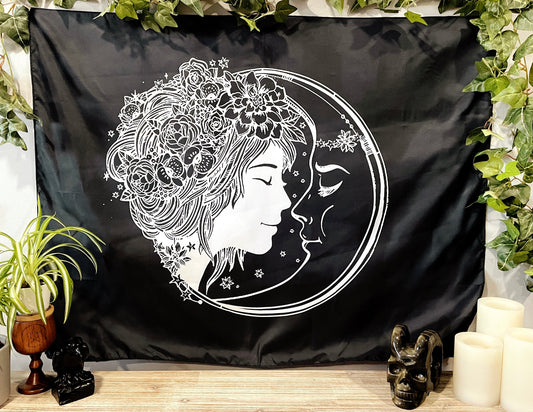 Pictured is a large wall tapestry with a black background and a woman with flowers in her hair smiling at a crescent moon printed in white on it. Moon Lover Wall Tapestry 