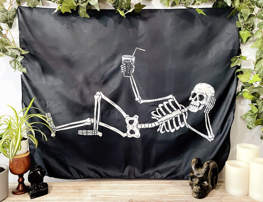 Pictured is a large wall tapestry with a black background and a large skeleton lounging with a drink in its hand printed in white on it. Relaxing Skeleton Wall Tapestry 