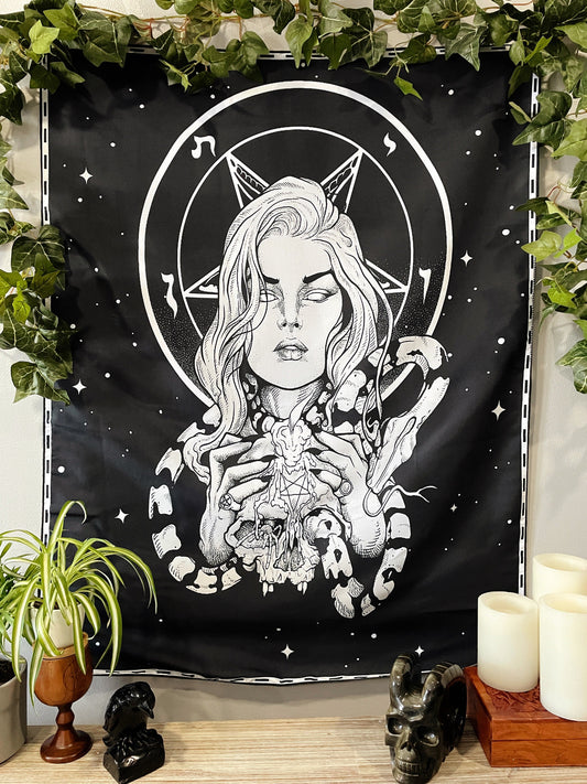 Pictured is a large wall tapestry with a black background and a woman holding a skull with a snake wrapped around it printed in white on it. Woman & Snakes Wall Tapestry