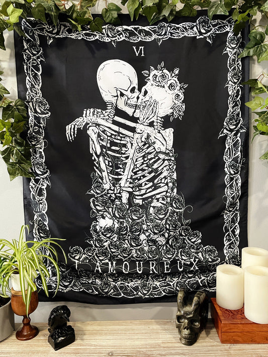 Pictured is a large wall tapestry with a black background and two skeletons kissing on a tarot card "the lovers" printed in white on it. The Lovers Wall Tapestry