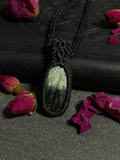 Pictured is a seraphinite cabochon wrapped in macrame thread. A gothic book and flowers are nearby. Seraphinite Macramé Necklace (Twisted Nightshade Jewellery) - The Wandering Fox Emporium, Your Crystal Store close up