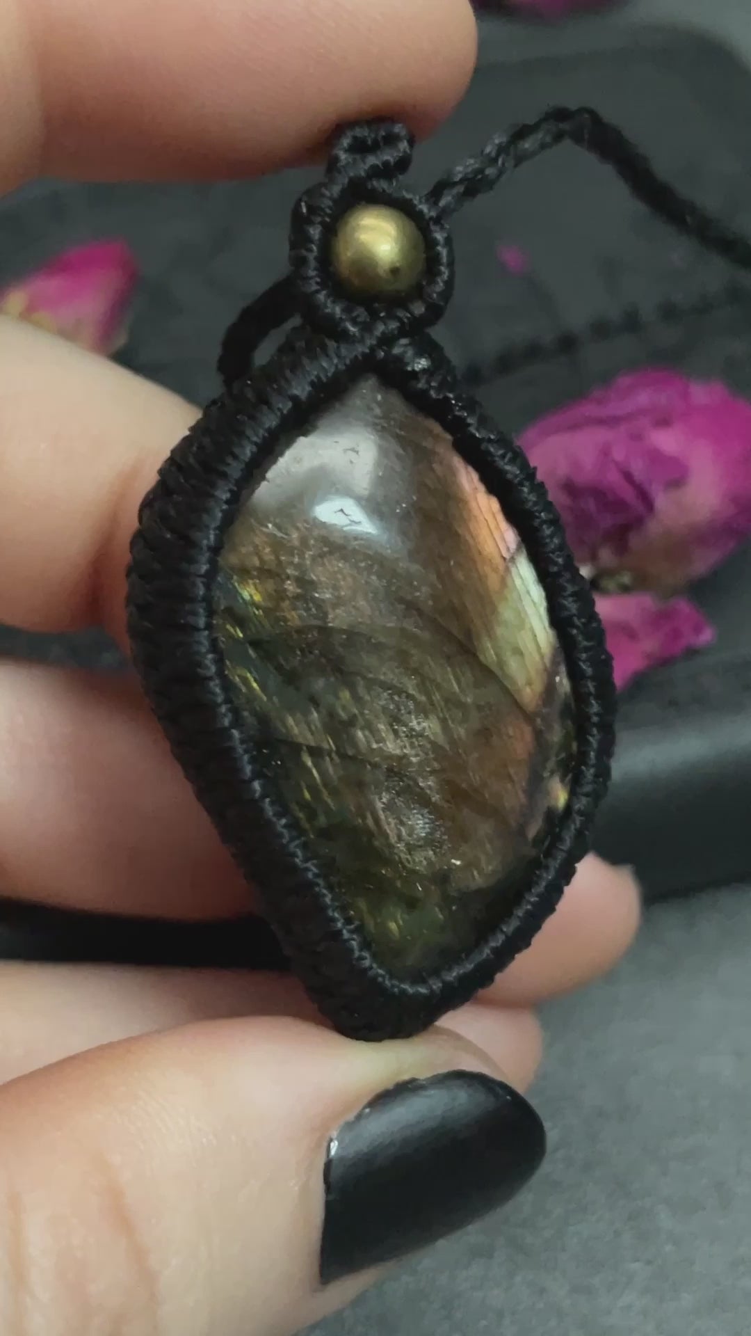 Pictured is a labradorite cabochon wrapped in macrame thread. A gothic book and flowers are nearby. Sunset Labradorite Macramé Necklace (Twisted Nightshade Jewellery) - The Wandering Fox Emporium, Your Crystal Store video